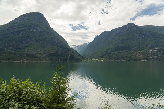 View of emerald waters reflecting the sky, framed by colossal green mountains under a sky of scattered clouds, nature's grandeur unfolds, Sorfjorden, Odda, Vestland, Norway.