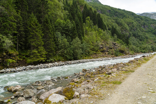 View of a rushing river, its turquoise waters contrasting with the rugged, rocky banks and the vibrant green forest clinging to the steep mountainside, Buarbreen, Odda, Norway.