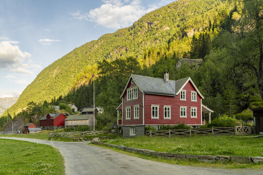 View of a quaint red house nestled against a towering green mountain, bathed in soft sunlight, beside a winding road, creating a serene landscape, buarbreen, Odda, Norway.