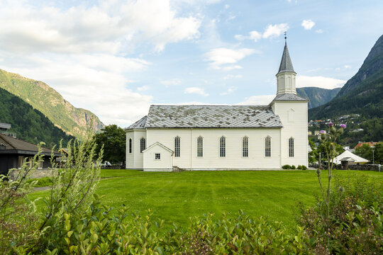 View of a white church with a grey roof and spire rises from a vibrant green lawn against a backdrop of towering mountains, Odda, Vestland, Norway.