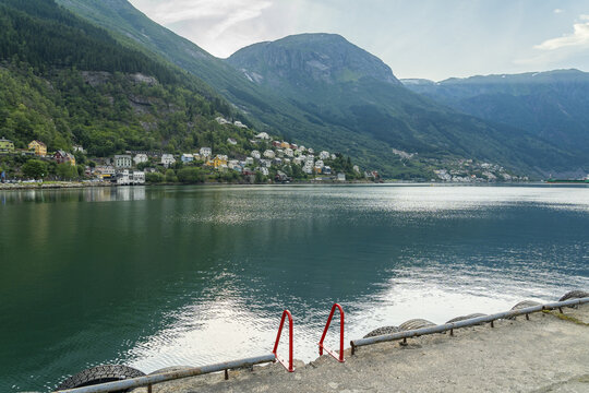 View of serene waters reflecting the sky, with a town nestled against verdant mountainsides, the scene framed by a concrete pier and metal ladder, Odda, Vestland, Norway.