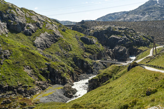 View of rugged, verdant hillsides embrace a rushing river beneath a dam of dark stone under a summer sky, Kvinnherad, Vestland, Norway.