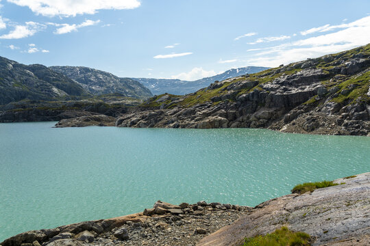 View of tranquil turquoise Mosevatnet waters reflecting the sky under scattered clouds, bordered by rugged hills and rocky terrain, Kvinnherad, Vestland, Norway.