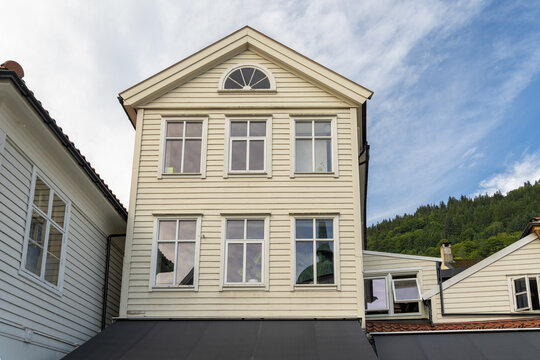 View of a charming, light-colored wooden house with a black awning stands tall against a backdrop of lush green trees and a partly cloudy sky, Bergen, Vestland, Norway.