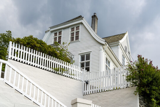 View of a stately white house with a quaint picket fence, nestled amongst verdant foliage under a dramatic sky, Bergen, Vestland, Norway.