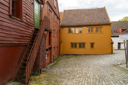 View of striking red and yellow wooden buildings rise against a cloudy sky, connected by an exterior staircase over cobblestone ground, Bergen, Norway.