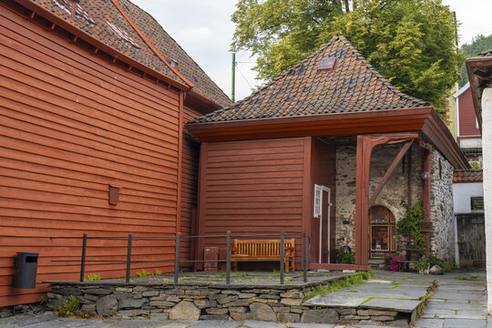 View of red wooden buildings with tiled roofs, stone foundations, and a bench, evoking a sense of history and charm, Bergen, Norway.