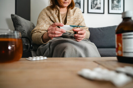 Person with flu checking fever on home couch with medicine and tea