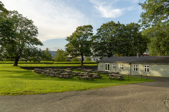 View of rows of wooden picnic tables nestled in a vibrant green lawn, framed by lush trees under a soft sky, Bergenhus fortress, Bergen, Vestland, Norway.