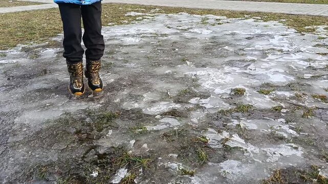 A child in black pants and black and yellow boots jumps in an icy puddle.