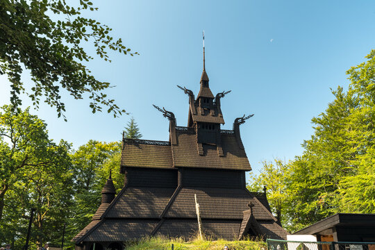 View of a dark, wooden Fantoft stave church, its intricate carvings reaching toward the clear sky, nestled amongst vibrant green trees, Bergen, Vestland, Norway.