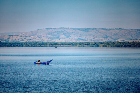 View of a vibrant blue boat cuts through the tranquil waters, a distant shoreline and rolling hills paint a serene backdrop, framed by a clear sky, Kampala, Uganda.