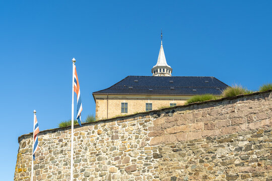 View of a historic stone fortress wall topped with green grass, flags waving against a clear blue sky, Oslo, Oslo, Norway.
