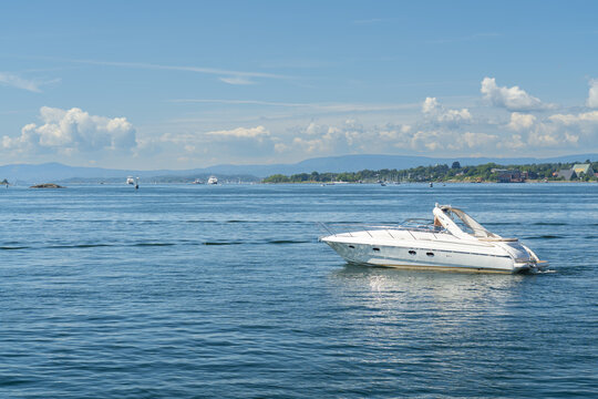 View of a sleek white motorboat glides across the shimmering blue water under a bright sky, with distant land a hazy green horizon, Oslo, Oslo, Norway.