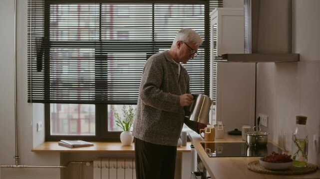 Side view medium long shot of senior Caucasian man in eyeglasses making drip coffee in modern kitchen, pouring hot water from electric kettle into two mugs and walking away