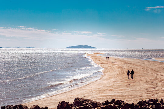 View of a couple walking hand-in-hand along a sandy strip as waves gently lap the shore under a clear blue sky, Seoul, South Korea.