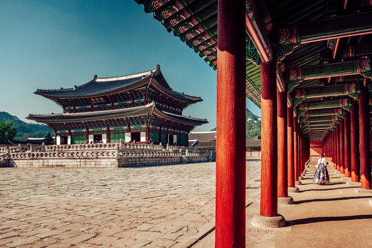 View of a woman in traditional dress walks through a corridor with vibrant red pillars, contrasting with the grand palace and expansive courtyard, Seoul, Seoul, South Korea.