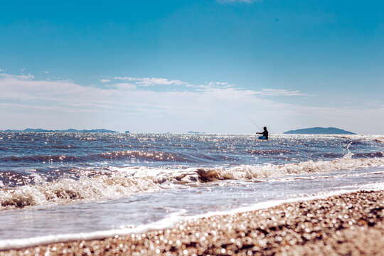View of a solitary figure fishing in the sea under a bright sky, with waves crashing on the sandy shore, creating a serene coastal scene, Seoul, South Korea.