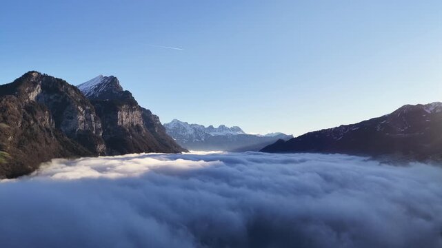Drone flying above thick cloud inversion between alpine cliffs revealing distant snow peaks and clear morning sky.