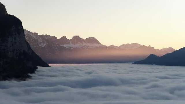 Drone sunset view over cloud covered Walensee valley with alpine silhouettes and warm twilight colors.