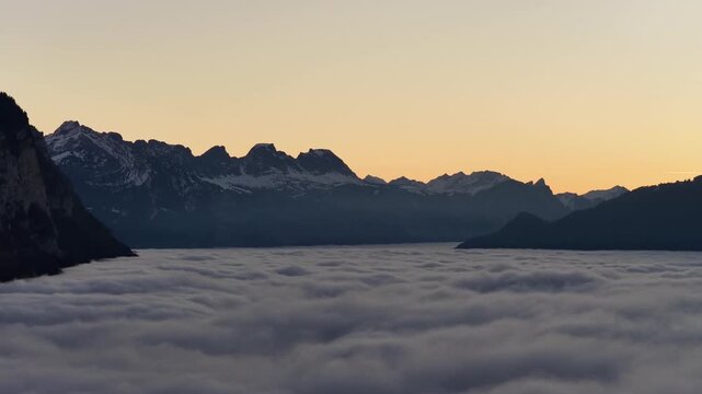 Lateral drone pan over cloud inversion at sunset revealing dramatic alpine silhouettes and glowing sky.