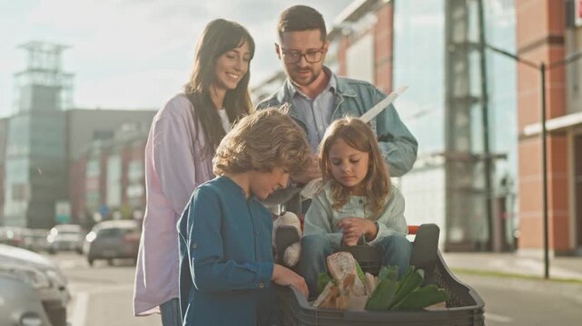 Family with two kids checking shopping receipt outside. Man holding bill, woman standing nearby, children looking at groceries in cart with bread and vegetables in parking lot after supermarket visit.