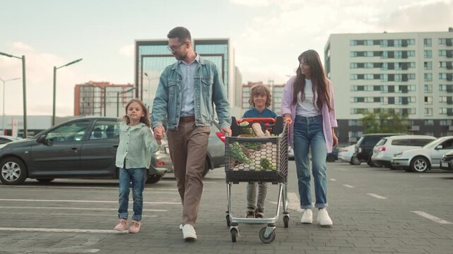 Family walking across parking lot after shopping. Father holding daughter&rsquo;s hand, son pushing cart full of groceries, mother smiling beside. Parents and children spending time together outdoors.