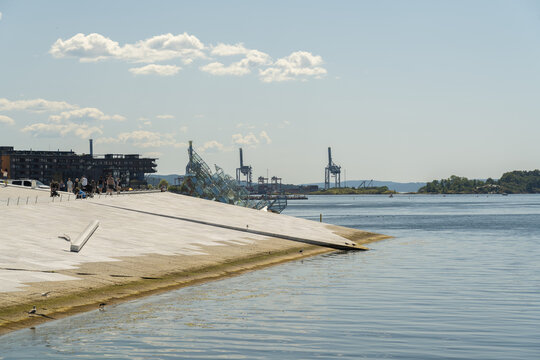 View of the serene waterfront where modern architecture meets the tranquil sea under a vast sky, with industrial cranes silhouetted against the horizon, Oslo, Oslo, Norway.