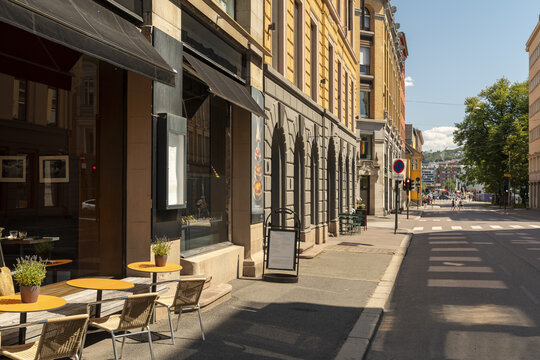 View of a quiet street with yellow tables and chairs outside a restaurant with black awnings, casting deep shadows on the sidewalk, Oslo, Oslo, Norway.
