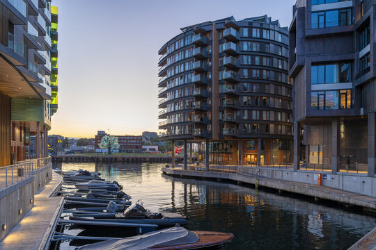 View of modern architecture reflected in the calm waters of a harbor, with boats gently swaying under a serene sky, Tjuvholmen, Oslo, Norway.