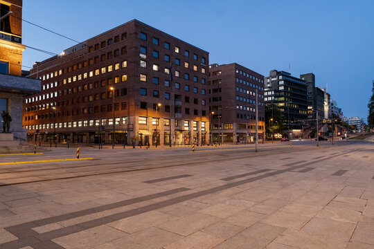 View of warm lights spill from modern buildings onto a patterned stone plaza under a cool blue twilight sky, at radhus plass square, Oslo, Norway.