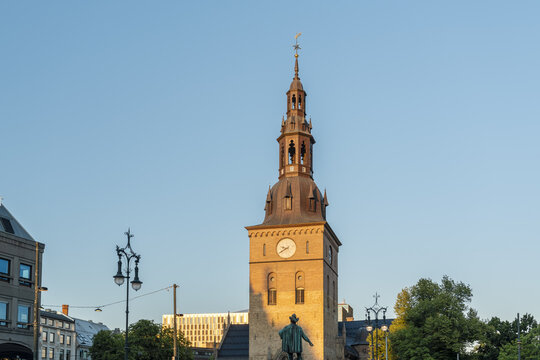 View of a tall, sunlit church tower with a clock face rising above a statue and green trees against a clear blue sky, Oslo, Oslo, Norway.