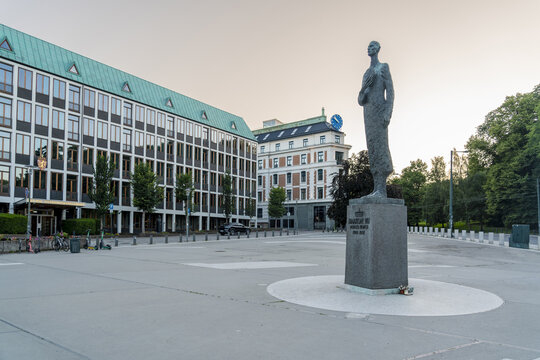 View of a bronze statue standing tall on a concrete base in a vast open square, framed by modern buildings against the soft glow of the sky, Oslo, Oslo, Norway.