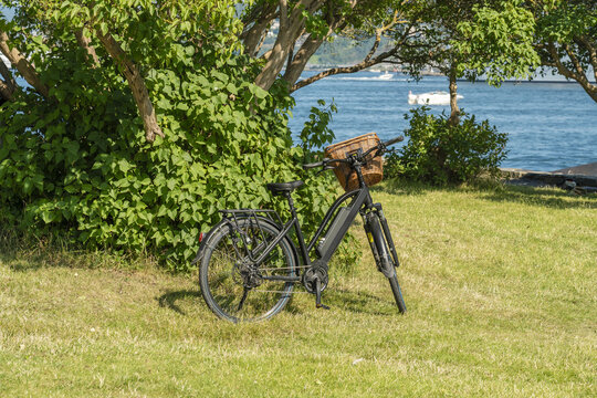 View of a bicycle rests on vibrant green grass, framed by lush trees against the shimmering blue of the water in the distance, Oslo, Oslo, Norway.