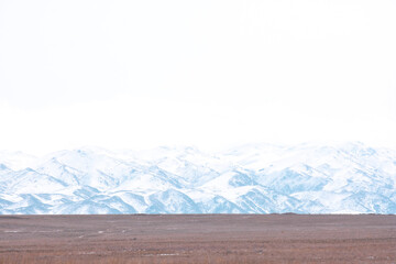 A vast snow-covered mountain range behind a flat brown field under a bright white sky. This minimalist high-key landscape is suitable for website headers, travel backgrounds, and environmental themes  © Lana Kray