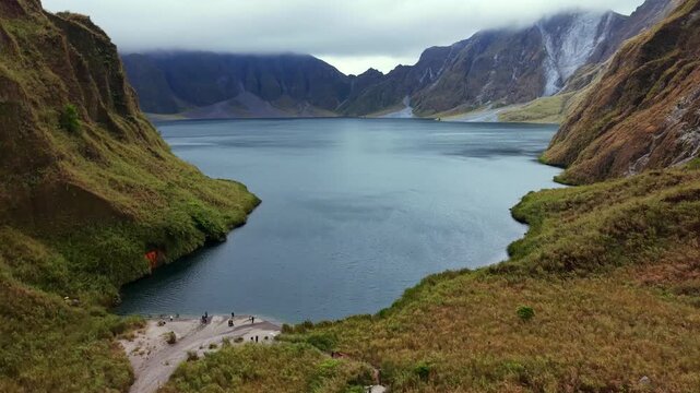 Serene mountain lake reflects overcast skies. Steep slopes frame the tranquil water. Hikers walk along the grassy path. They seem calm and content. Perfect for travel vlogs, nature documentaries