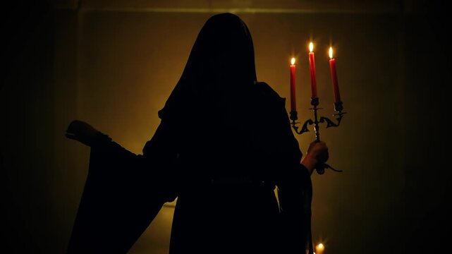 Silhouette fantasy victorian era woman in dark night black room holding old candlestick in hands candles burning warm light. Vintage retro style cape dress with hood on head. Sexy girl back rear view.