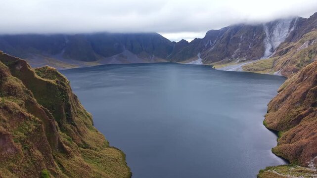 Pinatubo, philippines. Deep blue lake lies in a mountain valley. Misty peaks rise behind the water. Green hills slope down to the shore. Wide view shows calm waters reflecting the sky. 