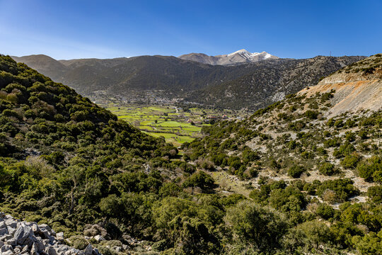 View of lush green valley nestled between rugged hills under a clear blue sky, with snow-capped mountains in the distance, Askifou, Chania, Greece.