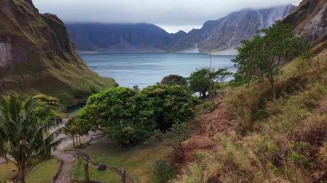 Lush green slopes frame a tranquil lake. Distant mountains rise under a cloudy sky. Wooden fence borders a winding stone path. Tropical trees line a grassy path. Water reflects the valley's calm hues