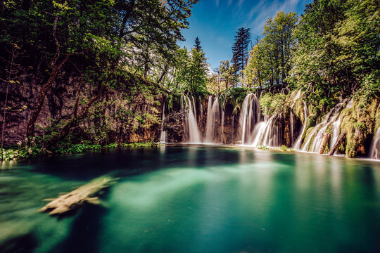 View of cascading waterfalls plunging into a serene, turquoise lake reflecting the vibrant green canopy of the surrounding forest, Plitvicka Jezera, Lika-Senj County, Croatia.