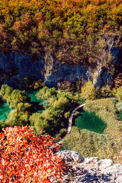 View of a wooden pathway snaking through vibrant turquoise waters, flanked by lush greenery and steep cliffs under autumn foliage, Plitvicka Jezera, Lika-Senj County, Croatia.
