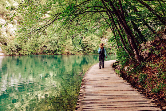 View of a solitary figure strolls along a wooden path, bordered by lush greenery and turquoise waters, creating a serene landscape, Plitvicka Jezera, Lika-Senj County, Croatia.