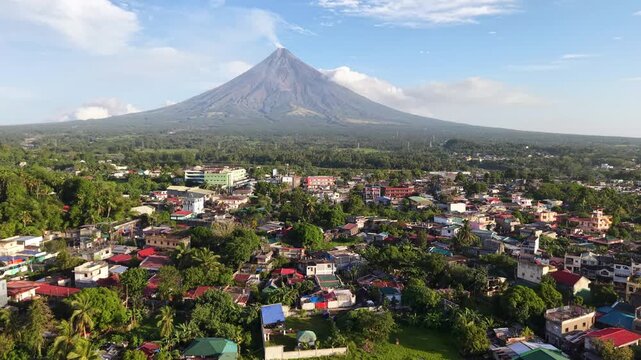 Mayon Volcano. Old stone church tower stands at the left. Town stretches with green fields and houses. Majestic volcano rises high in the distance. Blue sky with soft clouds. No people visible