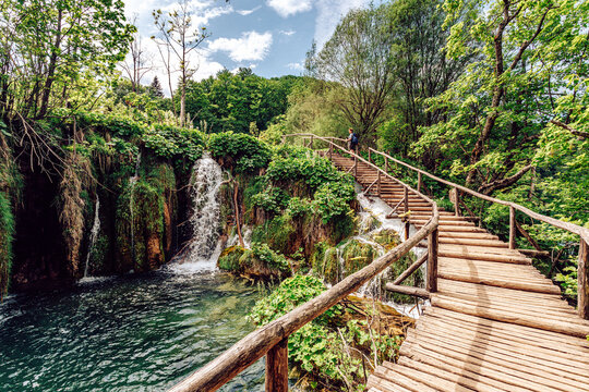 View of cascading waterfall plunges into a serene turquoise pool, framed by vibrant green foliage and rustic wooden pathways, Plitvicka Jezera, Lika-Senj County, Croatia.