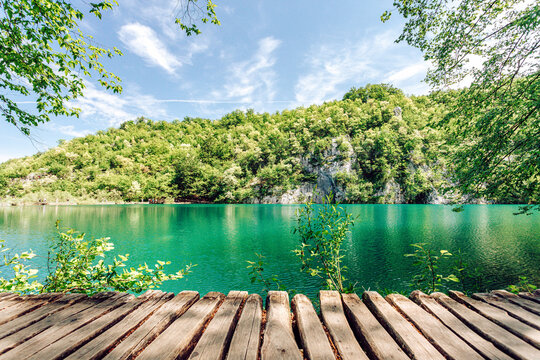 View of the turquoise lake waters reflecting the lush green forest hills, framed by a rustic wooden boardwalk, Plitvicka Jezera, Lika-Senj County, Croatia.