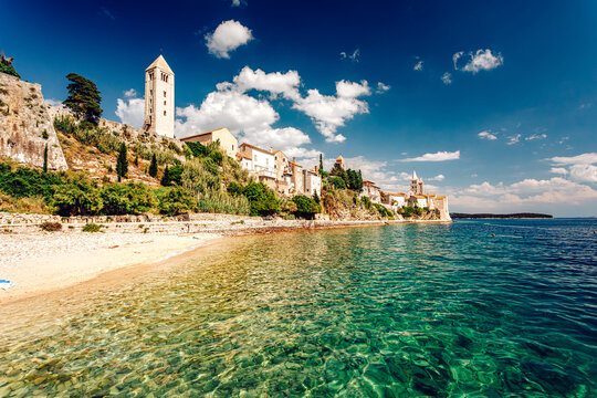 View of historic buildings perch atop rugged cliffs overlooking crystal-clear turquoise waters and a sandy beach, bathed in warm sunlight, Rab, Primorje-Gorski Kotar County, Croatia.