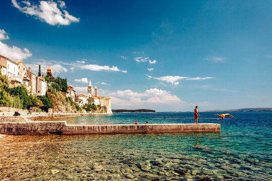 View of clear turquoise waters meet ancient stone buildings under a vibrant blue sky, as figures swim and dive off a pier, Rab, Primorje-Gorski Kotar County, Croatia.