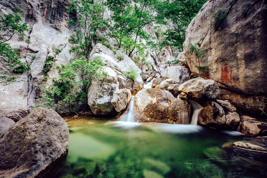 View of cascading waterfalls tumble over moss-covered rocks into a serene, emerald pool nestled amidst towering cliffs and vibrant green foliage, Starigrad, Zadar County, Croatia.