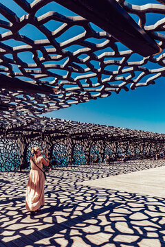 View of a woman stands beneath a modern, tessellated canopy, its geometric shadows dancing on the boardwalk under a clear blue sky, Marseille, Provence-Alpes-Cote d'Azur, France.